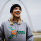 A woman standing in front of the St. Louis arch wearing a grey St. Patrick's Day St. Louis flag hoodie. 
