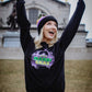A woman with her hands in the air wearing a black Soulard Mardi Gras sweatshirt standing in front of the Art Museum in St. Louis. 